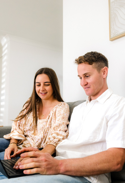 Two people sit on a grey couch, looking at a laptop together in a bright, modern living room.