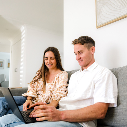 Two people sit on a grey couch, looking at a laptop together in a bright, modern living room.