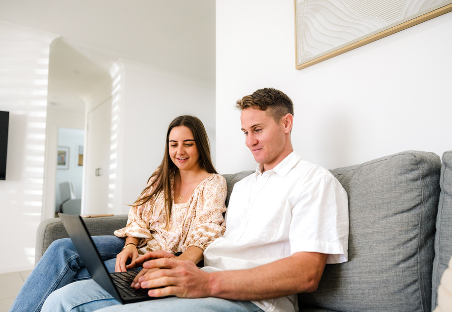 Two people sit on a grey couch, looking at a laptop together in a bright, modern living room.