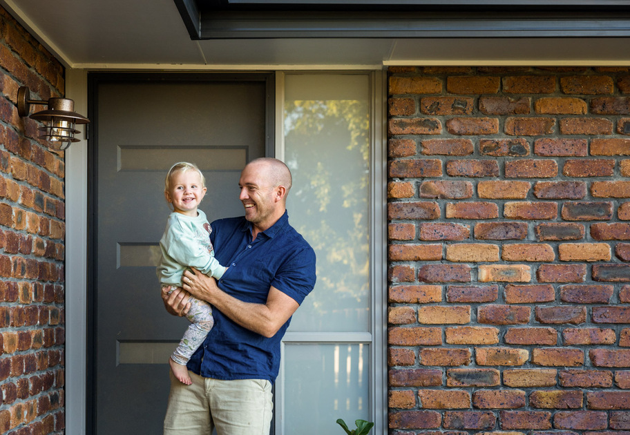 A man holding a smiling young child stands in front of a brick house near a gray door and window.
