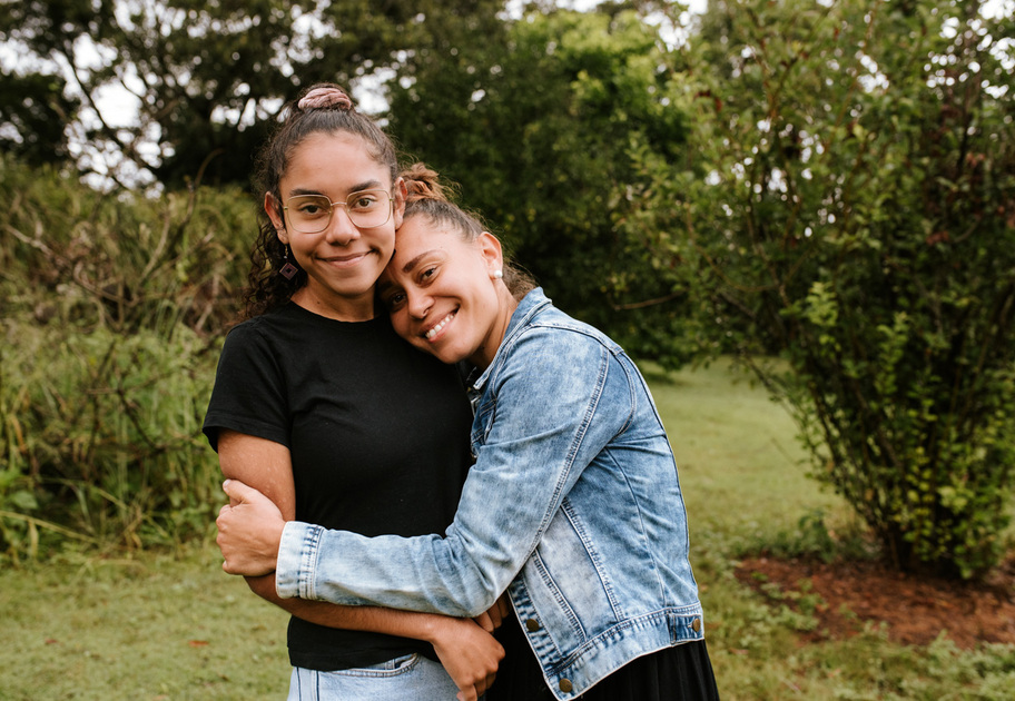Two women standing outdoors; one woman in a black shirt stands facing the camera while the other, in a denim jacket, hugs her and rests her head on her shoulder.