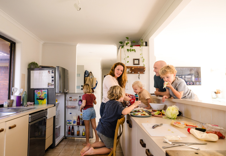 A family of five prepares food together in a bright kitchen, with children helping and parents smiling near a counter filled with ingredients.