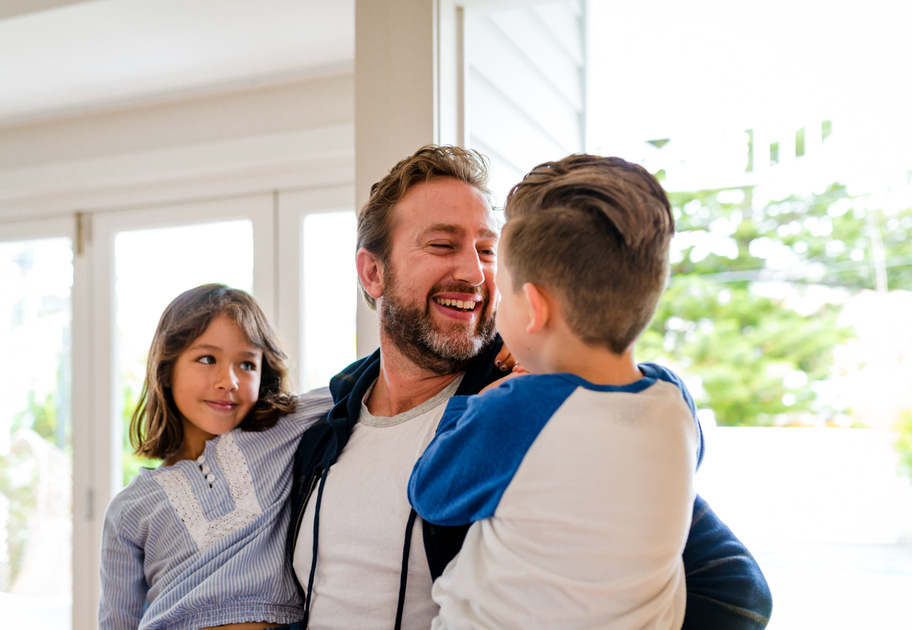 A bearded man smiling and holding a young boy and girl in his arms inside a bright room with large windows and greenery outside.