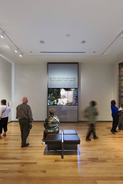 A woman sits on a bench observing the exhibit sign "Be not afraid, open wide the doors to Christ." A man stands to her left and a blurred woman walks to the right, showing the motion of the photograph.