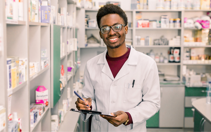 A smiling male medical professional wearing glasses and a white lab coat stands in a well-lit pharmacy aisle. He is holding a pen and clipboard, looking directly at the camera with shelves of medical supplies behind him.