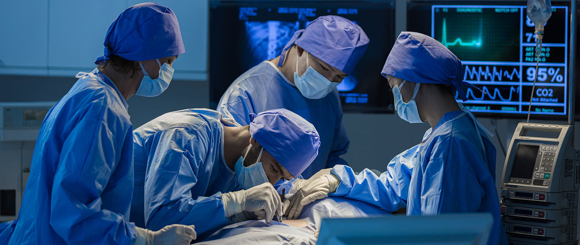 A medical team of four surgeons and nurses wearing blue surgical gowns, caps, masks, and gloves performs a procedure in a dimly lit operating room. Vital sign monitors and medical equipment are visible in the background.