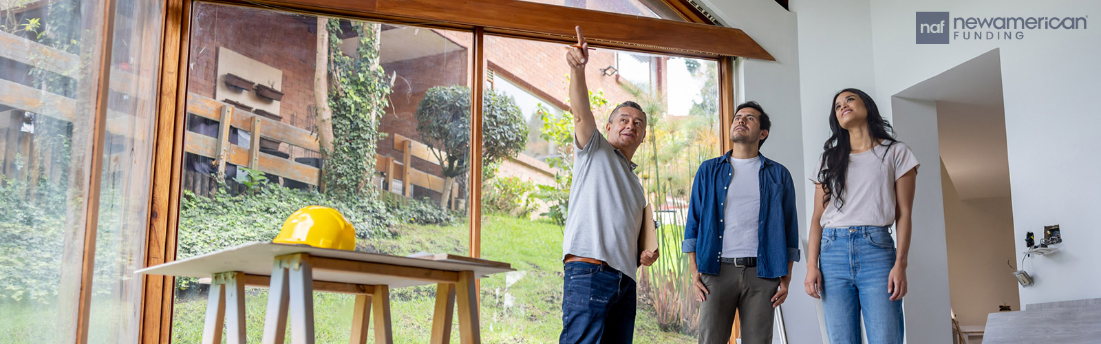An older Hispanic man and a younger Hispanic man and woman stand talking in a home that is under construction