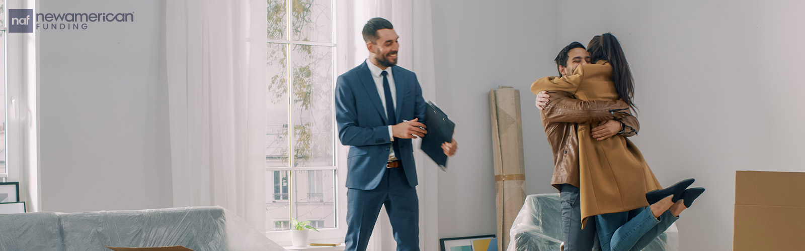 A smiling man pics a woman up in a hug inside of a home while a professional stands beside them smiling 