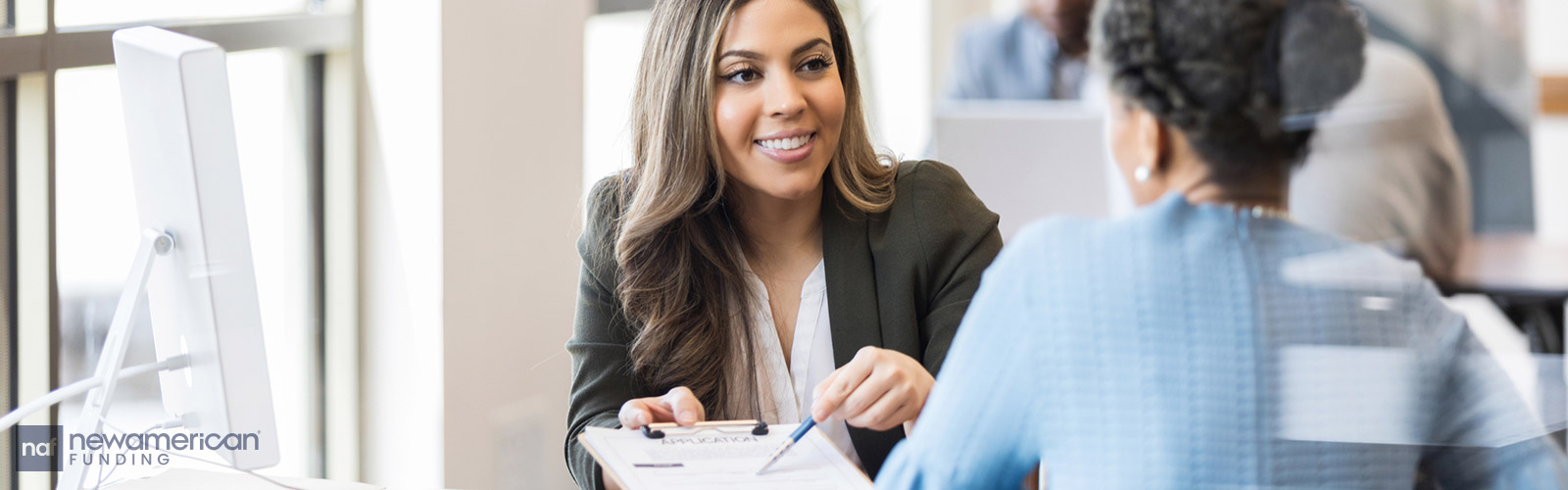 A woman dressed in a suit hands another woman a clipboard with paperwork on it