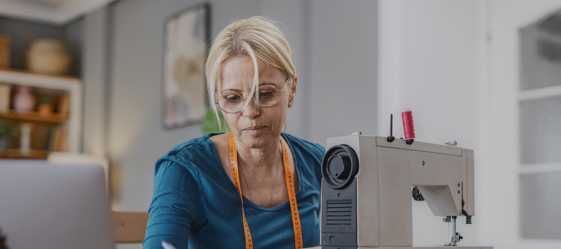 A woman writing something on a notebook while sitting in front of a sewing machine.