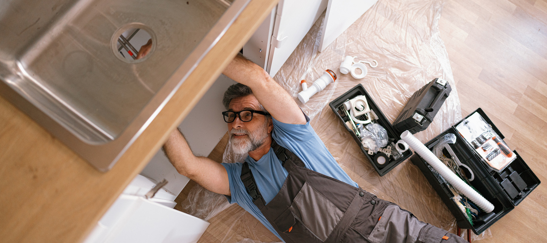 A man lying on the floor repairing pipes under a kitchen sink.