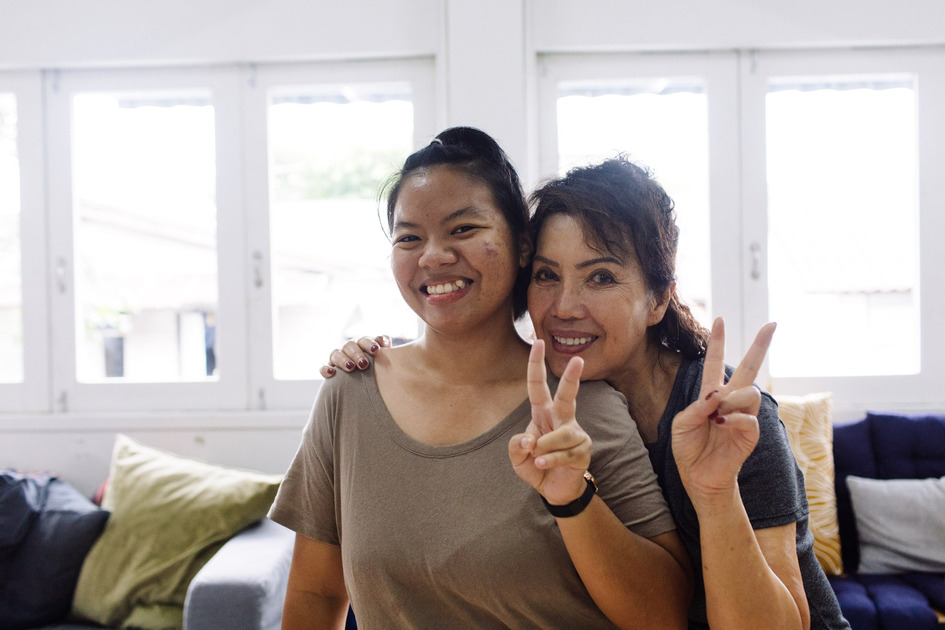 A daughter and her mum in the living room with their arms around each other, both smiling and making peace signs.