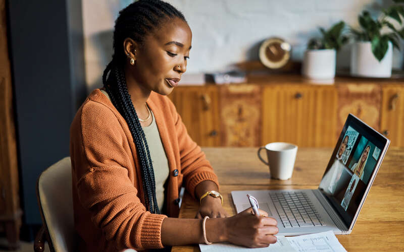 A woman sits at a desk writing. There are a laptop computer and a cup of coffee on the desk.