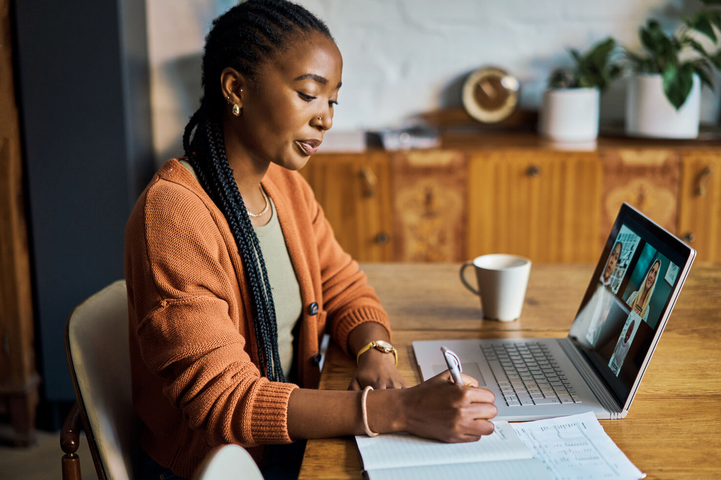 A woman sits at a desk writing. There are a laptop computer and a cup of coffee on the desk.