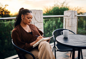 Woman sits in patio chair outside at sunrise or sunset journaling. She has a cup of coffee on the table next to her.