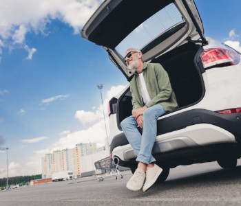 Moški sedi v prtljažniku svojega avtomobila in je zadovoljen, ker je uspešno znižal stroške zavarovanja svojega avta.