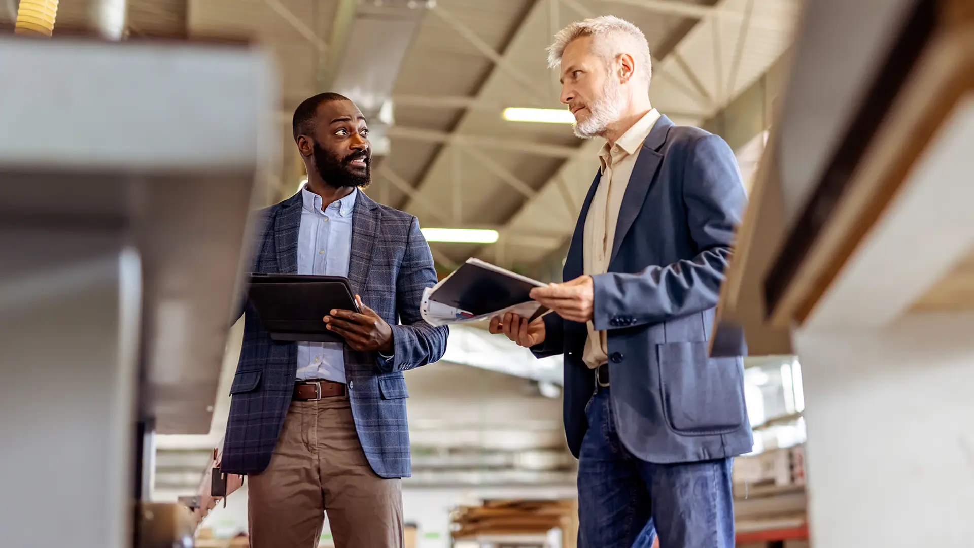 Two professionals discussing work in a modern factory while holding documents and a tablet.