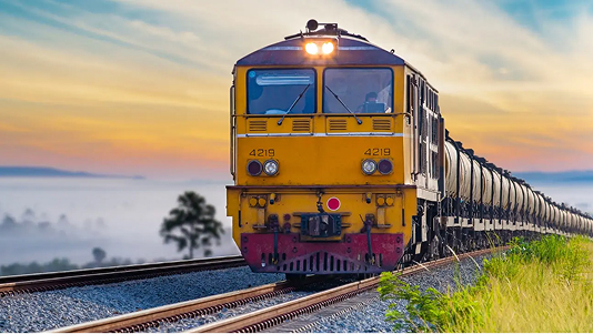 A long freight train with a yellow and red locomotive pulls a string of black tanker cars along a track. The train is moving through a grassy, rural landscape at sunrise or sunset, with a misty, colorful sky and rolling fog in the distance.