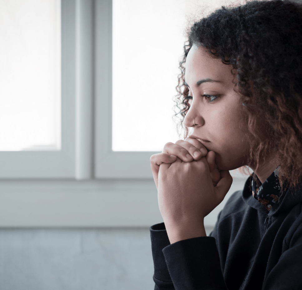 Young woman sitting indoors near a window, looking thoughtful with her hands clasped under her chin.