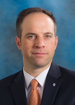 Headshot of Michael Votto, who smiles while wearing a black suit and a burnt orange tie