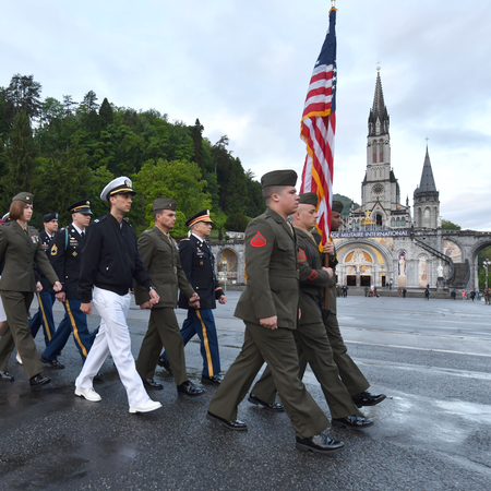 Various ranks of the miltary march at the Warriors to Lourdes Pilgimmage in front of the Basilica of Our Lady of the Immaculate Conception, men in the front hold an American flag.. 