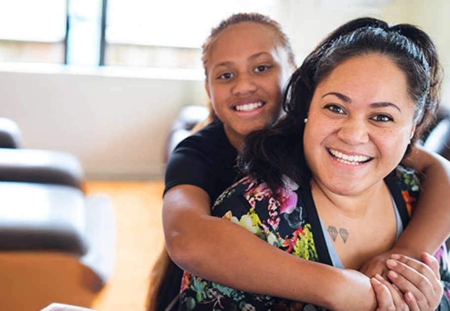 Sally smiling with a young girl hugging her from behind, both at home, featured in Mission Australia's Summer Impact Report 2025.