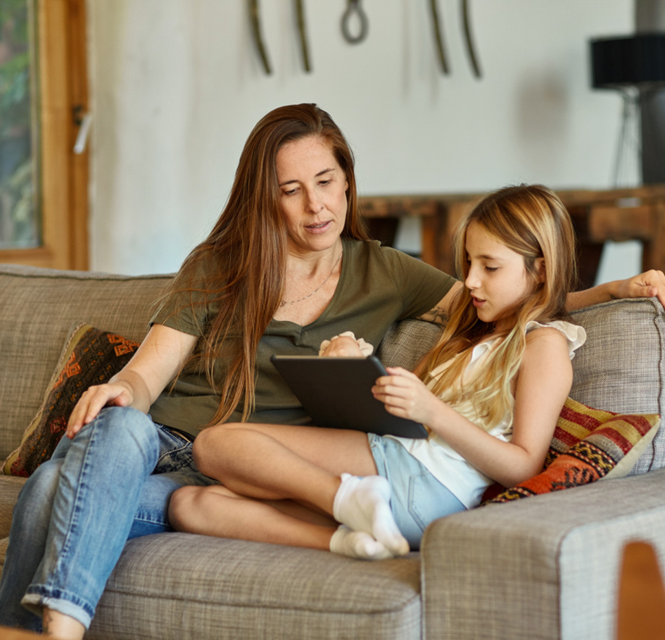A woman and a young girl sit together on a couch at home, looking at a tablet. The woman has her arm around the girl, and both appear focused on the screen. The setting is cozy and casual.