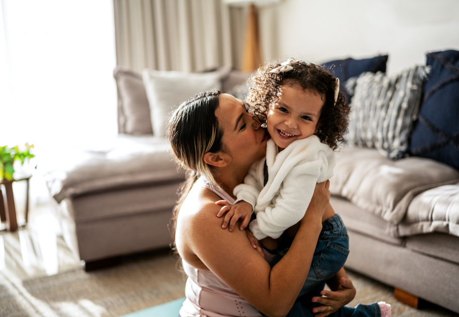 A woman kneels on the floor, hugging and kissing a smiling young child with curly hair in a cozy living room with a sofa and decorative pillows in the background.