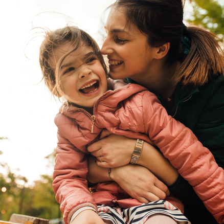 A woman embraces a smiling young girl outdoors on a sunny day. Both are wearing jackets and appear happy, sharing a joyful moment together with trees in the blurred background.
