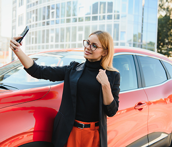 Mlada ženska ponosno pozira za sebek ob vzorno bočno parkiranem vozilu.