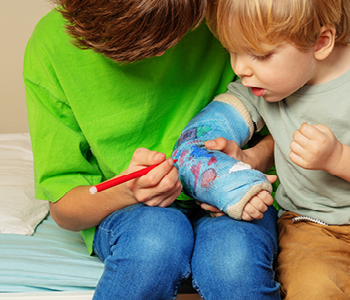Starejši deček mlajšemu riše na mavec na roki, medtem ko ga mlajši pozorno opazuje. Konec poškodb pri otrocih.