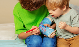 Starejši deček mlajšemu riše na mavec na roki, medtem ko ga mlajši pozorno opazuje. Konec poškodb pri otrocih.