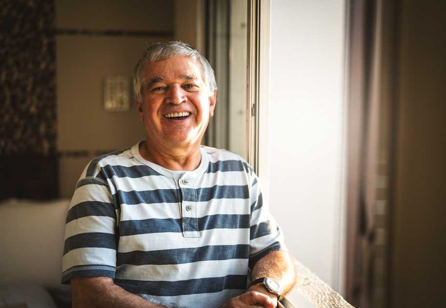An older man with gray hair, wearing a striped shirt, smiles while standing by a sunlit window indoors. The background is softly blurred, creating a warm and cheerful atmosphere.