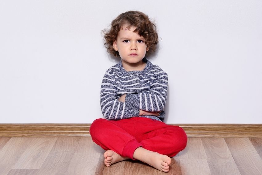Kodrolas deček sedi na tleh s prekrižanimi nogami in prekrižanimi rokami kujavo zre v kamero.