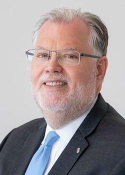 A corporate headshot style portrait of Peter Gervais, smiling wearing a dark suit, white shirt, and blue tie on a light grey background.