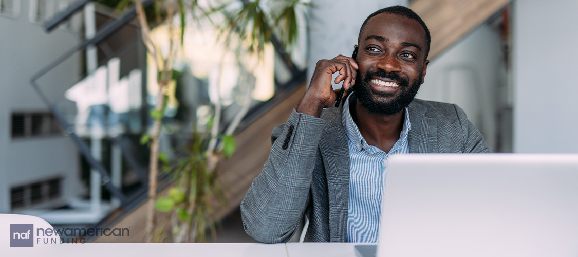 A man sitting in front of a laptop, talking on his cell phone, and smiling.