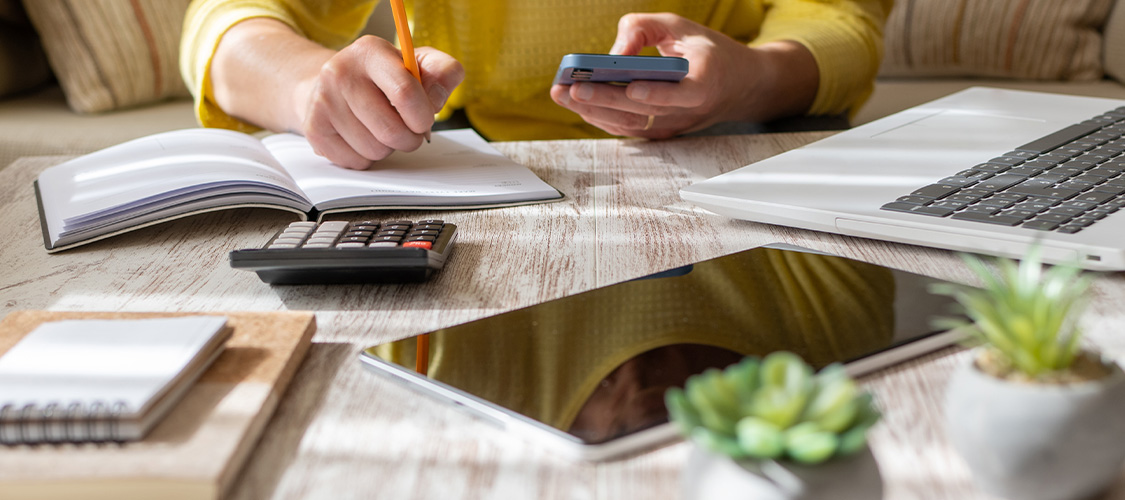 Someone writing in a journal while holding a phone with a calculator and tablet nearby.