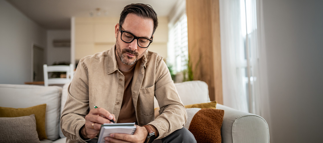 A man wearing glasses sitting on a couch writing on a pad of paper.