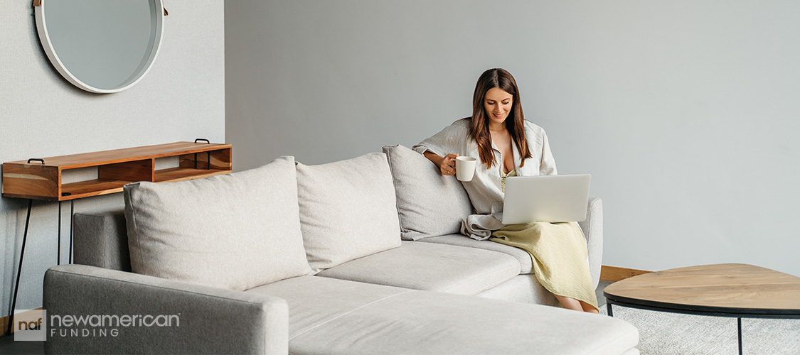 A woman sitting on a white couch in a light, uncluttered, grey room.