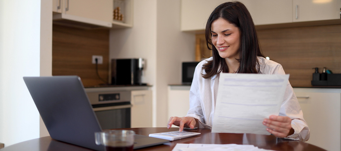 A woman on a laptop looking at paperwork at the kitchen table.