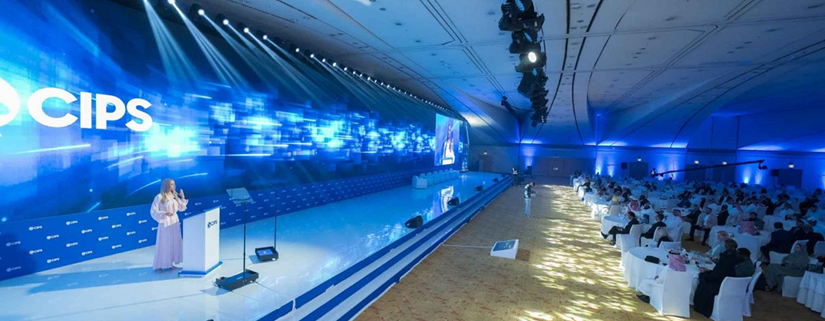 Wide view of a CIPS conference with a large stage, blue digital background, and audience seated at round tables.