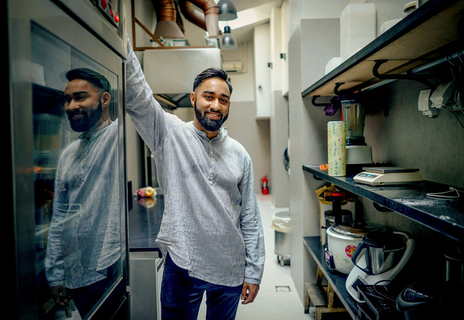 A man with a beard and short hair, wearing a light gray shirt, stands smiling in a modern kitchen. He leans against a tall appliance, with various kitchen equipment visible around him.