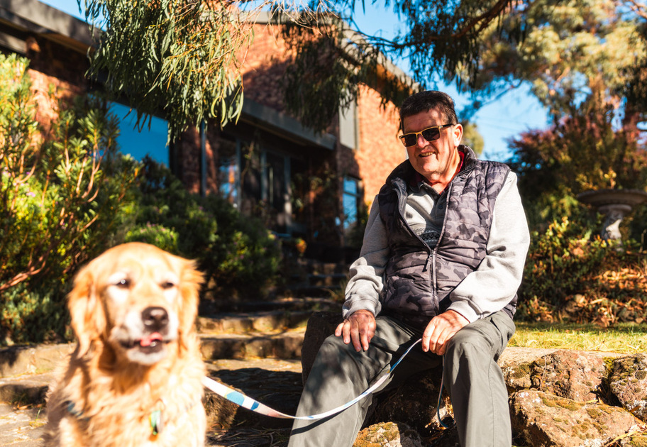 An older man wearing sunglasses sits on outdoor stone steps holding the leash of a golden retriever dog on a sunny day.