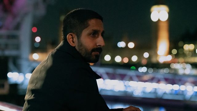 Sagar Radia as Rishi Ramdani leaning on a bridge railing at night, city lights glowing behind him.