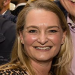 A close-up portrait of a smiling woman with blonde, shoulder-length hair. She is wearing a leopard-print blouse and a gold necklace, appearing to be at a social gathering with others partially visible in the blurred background.