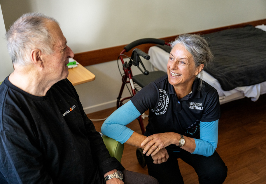 	
A smiling woman in a Mission Australia shirt kneels beside an elderly man sitting in a chair, engaging him in conversation in a cozy, well-lit room with a bed and a walker in the background.