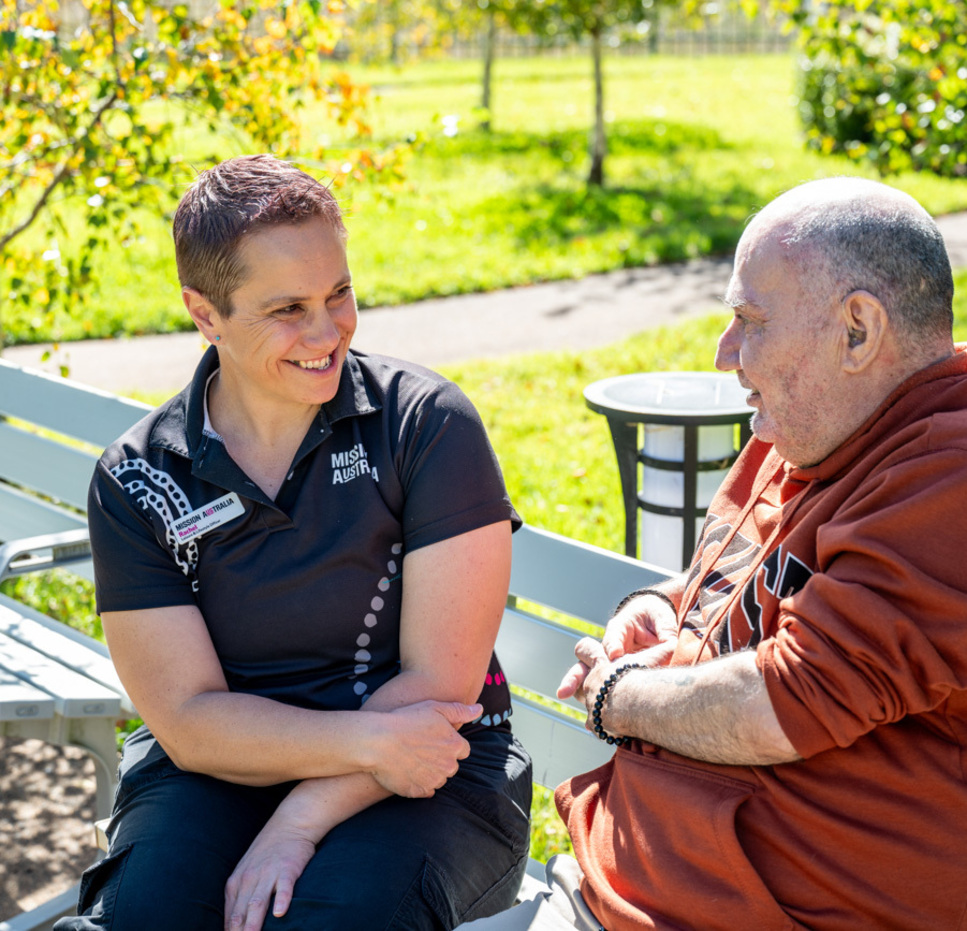 A woman and an older man sit on a park bench, smiling and talking together on a sunny day, surrounded by green grass and trees.