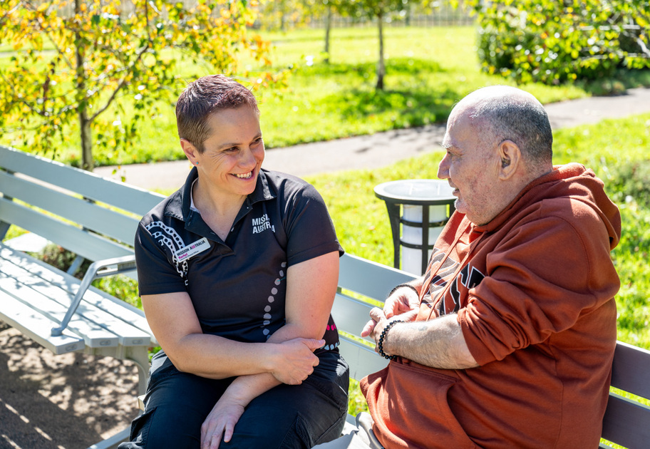 A woman and an older man sit on a park bench, smiling and talking together on a sunny day, surrounded by green grass and trees.