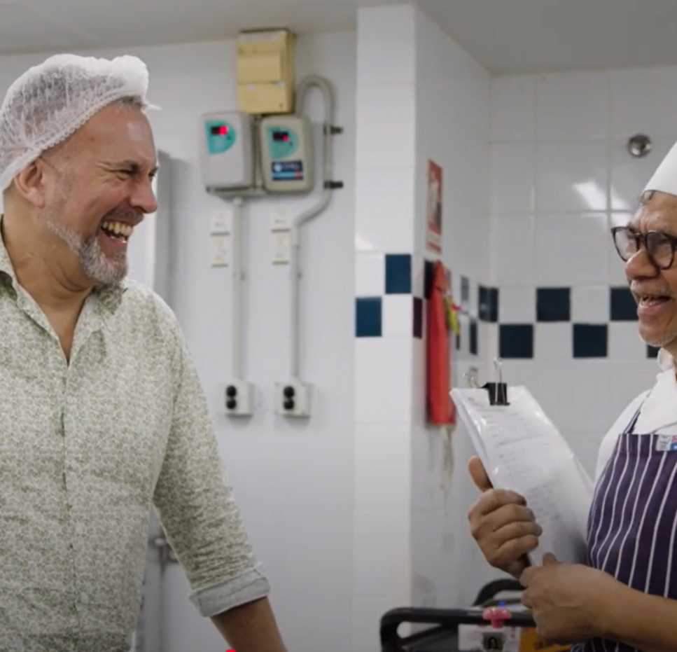 Two men smiling and laughing together in an aged care kitchen, one wearing a chef’s hat and apron with a clipboard, the other in a hairnet and patterned shirt.
