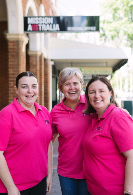Three women wearing Mission Australia shirts smile while standing together on a city sidewalk in front of a brick building with a Mission Australia Regional Office sign.